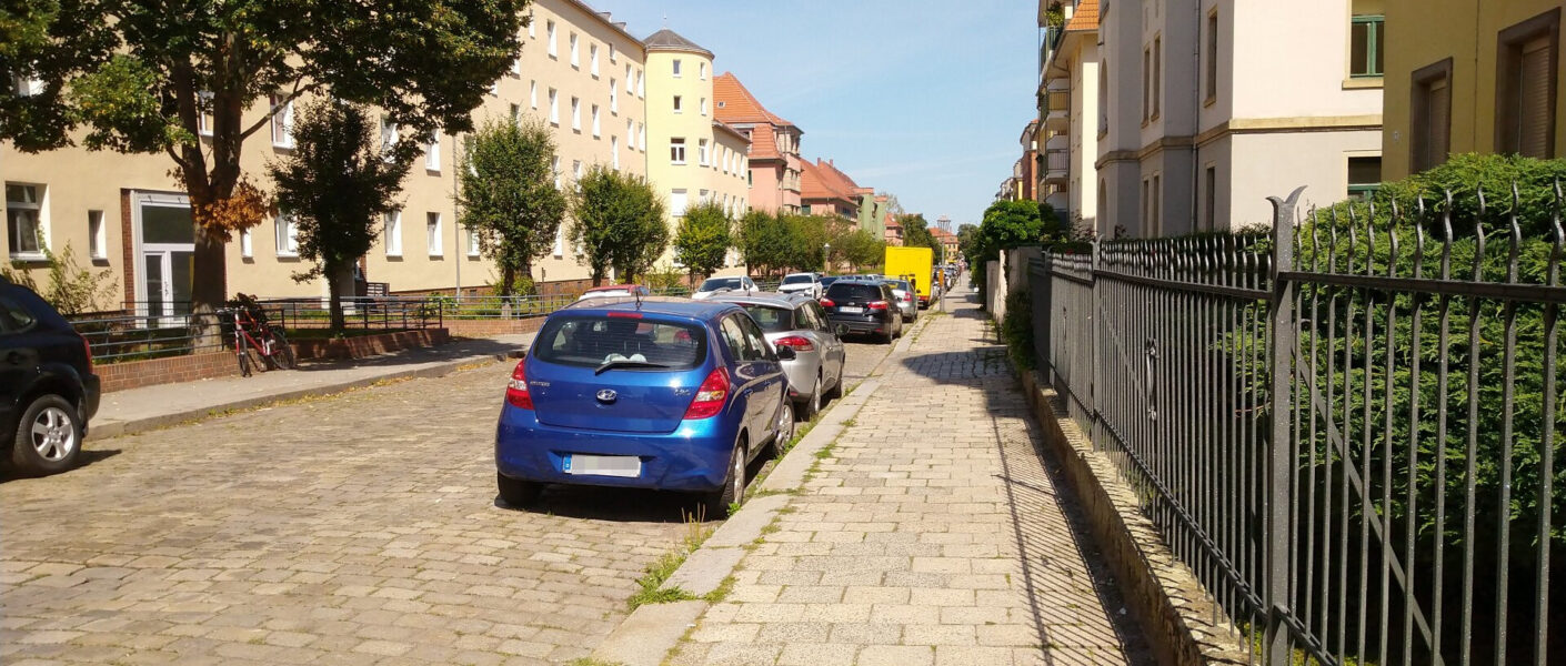 Eine Straße in der Sonne, rechts und links Häuser, je ein Fußweg und parkende Autos und freie Parkplätze dazwischen.