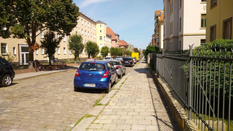 Eine Straße in der Sonne, rechts und links Häuser, je ein Fußweg und parkende Autos und freie Parkplätze dazwischen.