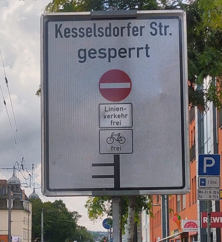 Ein Verkehrsschild mit schwarzen Balken für die betroffenen Straßen. Unter dem Text "Kesselsdorfer Str. gesperrt" ein dicker, schwarzer Balken, der die Kesselsdorfer symbolisiert. Oben am Balken ein rotes Einbahnstraßenschild, darunter "Linienverkehr frei" und Fahrrad frei. Nach links gehen zwei kurze, schwarze Striche für die Post- und Bünaustraße.