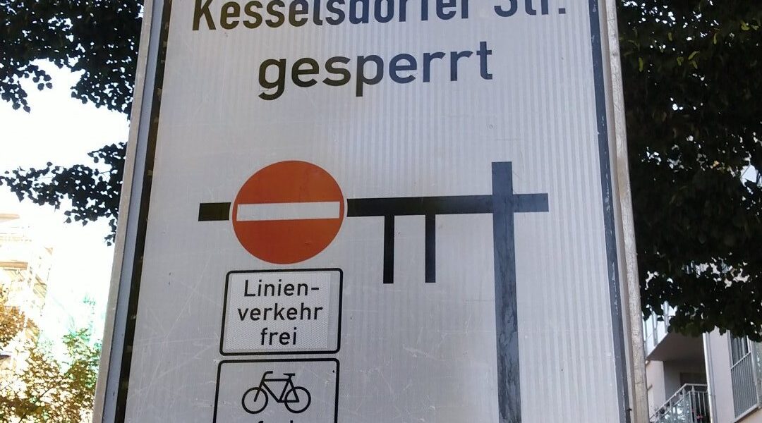 Ausschnitt eines Verkehrsschilds mit schwarzen Balken für die betroffenen Straßen. Links ein rotes Einbahnstraßenschild, darunter "Linienverkehr frei" und Fahrrad frei. Über den Balken die Schrift "Kesselsdorfer Str. gesperrt".