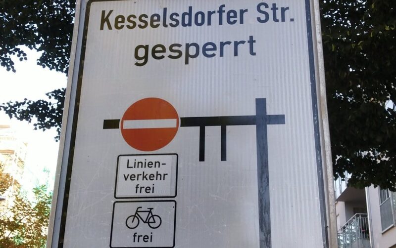 Ausschnitt eines Verkehrsschilds mit schwarzen Balken für die betroffenen Straßen. Links ein rotes Einbahnstraßenschild, darunter "Linienverkehr frei" und Fahrrad frei. Über den Balken die Schrift "Kesselsdorfer Str. gesperrt".